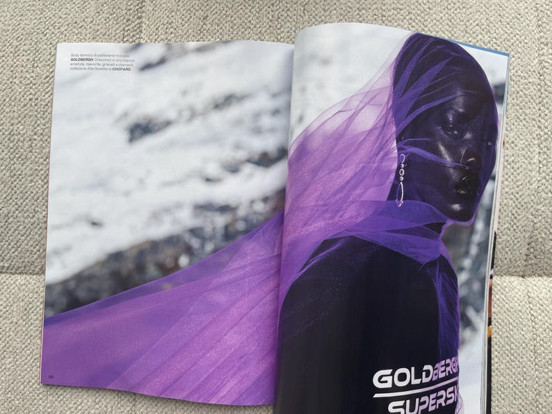 Puede incluir: Una p&aacute;gina de revista de moda con una persona de piel oscura, envuelta en una tela p&uacute;rpura transparente y fluida. La persona lleva pendientes plateados. El t&iacute;tulo de la revista, "GOLDBERG SUPERS", es visible en la parte inferior.