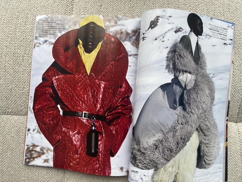 Puede incluir: Una revista de moda con dos modelos en atuendos de invierno. Una modelo lleva un abrigo rojo texturizado con cintur&oacute;n negro y un accesorio negro. La otra modelo lleva un abrigo de piel gris y un atuendo blanco, sobre un fondo nevado.