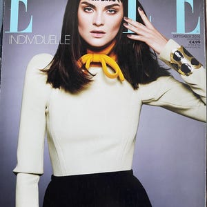 May include: Cover of Elle magazine featuring a woman with dark hair and bangs. She wears a cream-colored top with a yellow scarf and a black skirt. The magazine title "ELLE" is visible, along with the word "INDIVIDUELLE".
