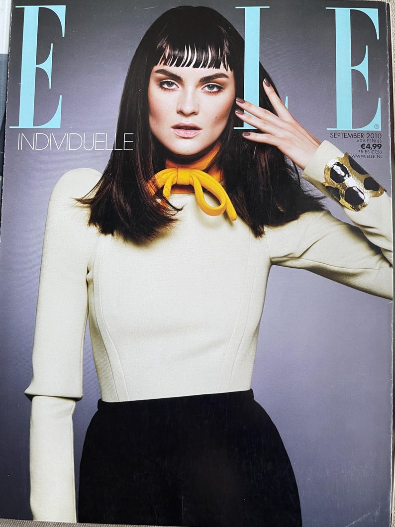 May include: Cover of Elle magazine featuring a woman with dark hair and bangs. She wears a cream-colored top with a yellow scarf and a black skirt. The magazine title "ELLE" is visible, along with the word "INDIVIDUELLE".