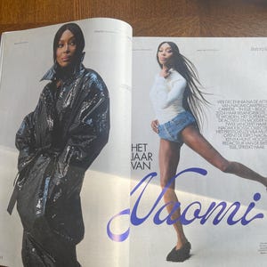 May include: A magazine spread with Naomi Campbell. One image shows her in a black, shiny coat, the other in a white top and denim shorts. The text "Het Jaar Van Naomi" is displayed in a stylized font.