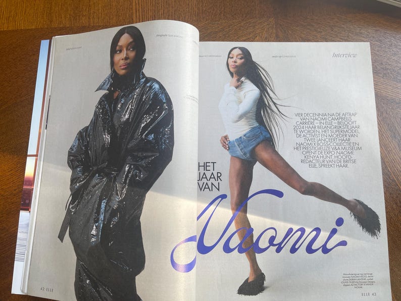 May include: A magazine spread with Naomi Campbell. One image shows her in a black, shiny coat, the other in a white top and denim shorts. The text "Het Jaar Van Naomi" is displayed in a stylized font.