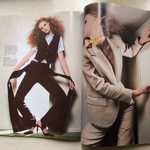 May include: Two women wearing different styles of clothing. The woman on the left is wearing a black pantsuit with a white vest and a long black scarf. The woman on the right is wearing a beige blazer with a gold bow tie and a pair of beige pants.
