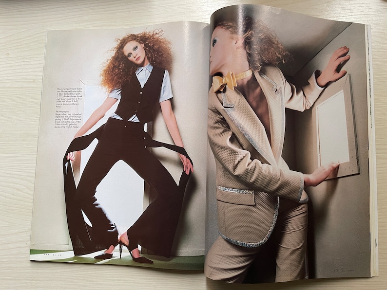 May include: Two women wearing different styles of clothing. The woman on the left is wearing a black pantsuit with a white vest and a long black scarf. The woman on the right is wearing a beige blazer with a gold bow tie and a pair of beige pants.