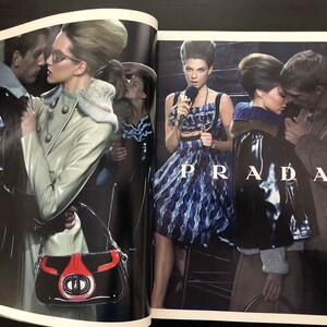 以下が含まれることがあります： 雑誌の2ページにわたるファッションエディトリアルで、モデルが革のジャケット、ドレス、白いコートを着ています。モデルは暗い工業的な場所でポーズをとっています。ページの下部に「PRADA」という文字が大きく印刷されています。