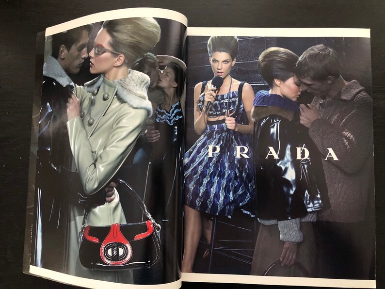 以下が含まれることがあります： 雑誌の2ページにわたるファッションエディトリアルで、モデルが革のジャケット、ドレス、白いコートを着ています。モデルは暗い工業的な場所でポーズをとっています。ページの下部に「PRADA」という文字が大きく印刷されています。