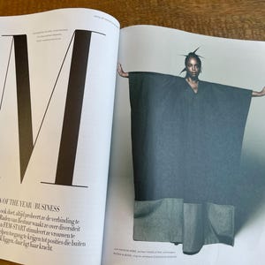 May include: A fashion magazine spread featuring a model wearing a dark gray cape-like garment with large pockets. The model is positioned with arms outstretched. The left page displays a large letter "M" and text that reads "WOMEN OF THE YEAR BUSINESS".