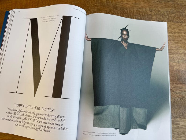May include: A fashion magazine spread featuring a model wearing a dark gray cape-like garment with large pockets. The model is positioned with arms outstretched. The left page displays a large letter "M" and text that reads "WOMEN OF THE YEAR BUSINESS".