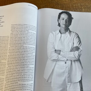 May include: Black and white photograph of a woman with short hair, wearing a white shirt and pants, with her arms crossed. The image is from a magazine, with text visible on the open pages.