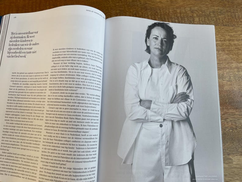 May include: Black and white photograph of a woman with short hair, wearing a white shirt and pants, with her arms crossed. The image is from a magazine, with text visible on the open pages.