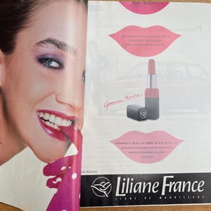 May include: An advertisement for Liliane France makeup. The image features a woman with bright pink lipstick and eye makeup. A lipstick tube and a small black compact are also shown. The text "Liliane France" is at the bottom.