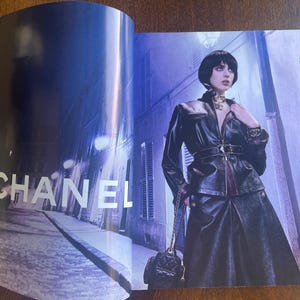 May include: A fashion magazine spread featuring the CHANEL logo in white. A person is wearing a black leather jacket, skirt, and carrying a quilted black purse. The background is a nighttime street scene with blue and purple hues.