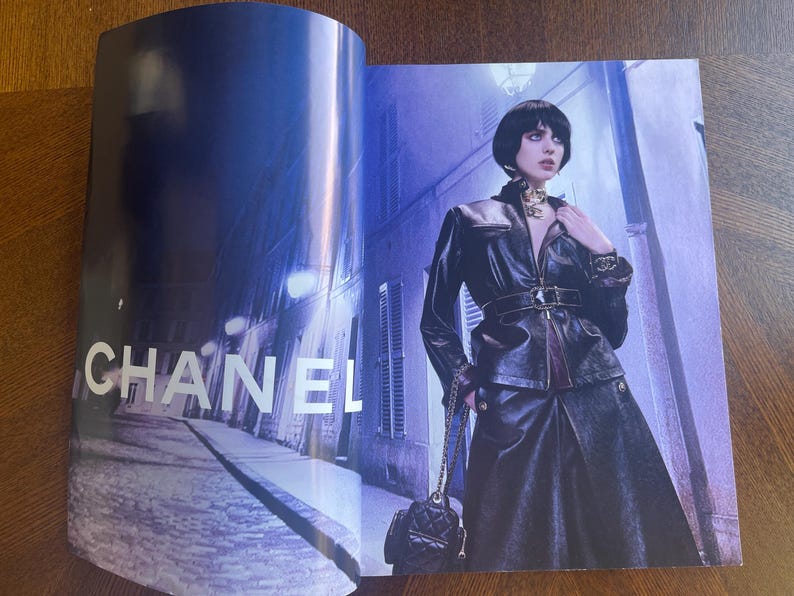 May include: A fashion magazine spread featuring the CHANEL logo in white. A person is wearing a black leather jacket, skirt, and carrying a quilted black purse. The background is a nighttime street scene with blue and purple hues.