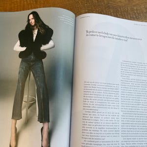May include: A fashion editorial spread featuring a person wearing a black top with a unique design, blue jeans, and high heels. The image is set against a neutral background. Text in the image reads, "Ik probeer met behulp van onze kunstwerken mensen weer in contact te brengen met de intuitieve taal."