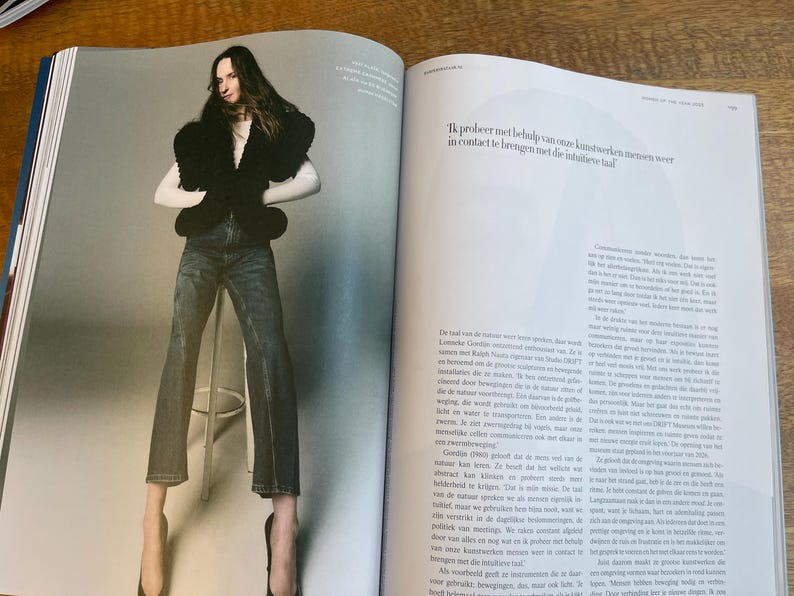 May include: A fashion editorial spread featuring a person wearing a black top with a unique design, blue jeans, and high heels. The image is set against a neutral background. Text in the image reads, "Ik probeer met behulp van onze kunstwerken mensen weer in contact te brengen met de intuitieve taal."