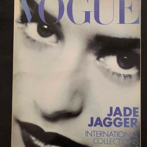 Op de afbeelding: Een vintage Vogue tijdschriftomslag met een zwart-wit close-up portret van Jade Jagger. De titel VOGUE staat in grote blauwe letters bovenaan. De tekst JADE JAGGER INTERNATIONAL COLLECTIONS ISSUE staat in het blauw rechtsonder.