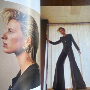 May include: A fashion editorial featuring a woman with blonde hair and a dark outfit. She wears a black jacket with gold buttons and wide-leg pants. The woman also wears a heart-shaped earring with a pearl design.