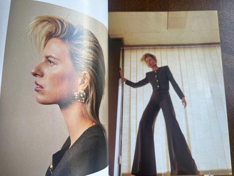 May include: A fashion editorial featuring a woman with blonde hair and a dark outfit. She wears a black jacket with gold buttons and wide-leg pants. The woman also wears a heart-shaped earring with a pearl design.