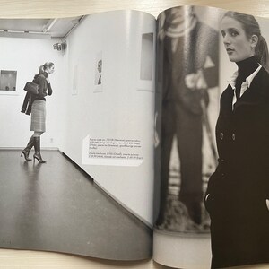 May include: A black and white photo of a woman wearing a black coat and a white turtleneck. She is standing in a room with a white wall and a large window. The woman is looking at the camera.