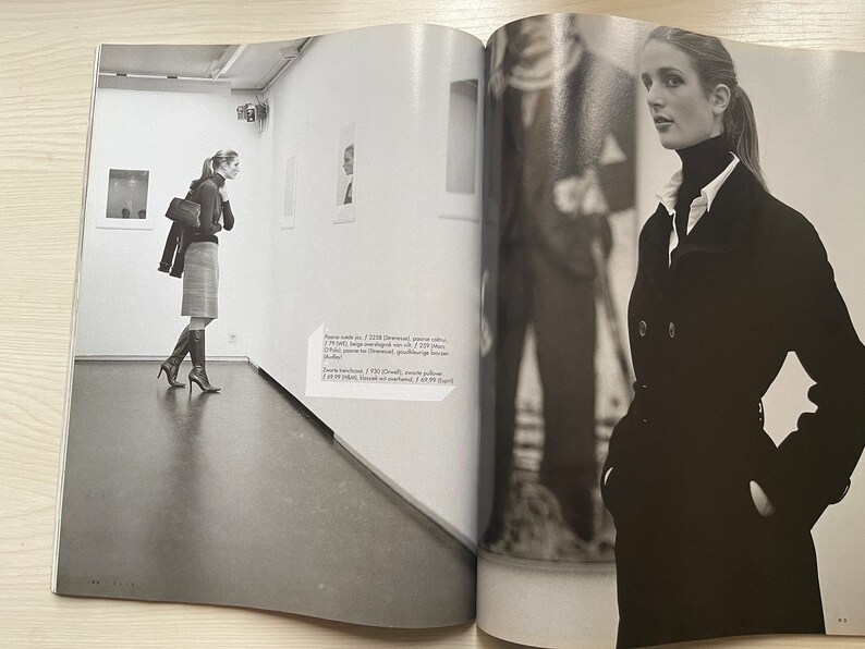 May include: A black and white photo of a woman wearing a black coat and a white turtleneck. She is standing in a room with a white wall and a large window. The woman is looking at the camera.