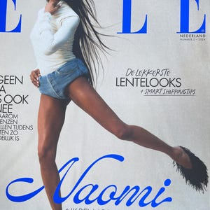 May include: Magazine cover of ELLE Nederland featuring Naomi Campbell. She wears a white top and denim shorts. The magazine title is in blue. Text includes "De lekkerste lentelooks + Smart shoppingtips" and "Ik ben absoluut niet perfect, maar ik doe wat ik kan."