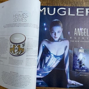 Puede incluir: Una doble p&aacute;gina de revista que presenta un anuncio de un nuevo perfume de Mugler llamado Angel Elixir. El anuncio presenta a una modelo con un atuendo met&aacute;lico plateado y un primer plano del frasco de perfume. El texto del anuncio dice "Angel Elixir La Nueva Fragancia" y "Mugler Recargable".