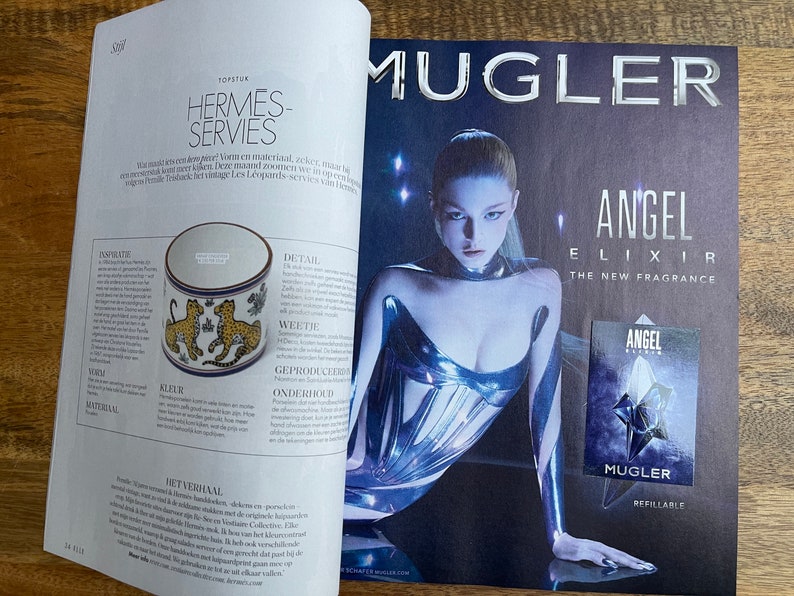 Puede incluir: Una doble p&aacute;gina de revista que presenta un anuncio de un nuevo perfume de Mugler llamado Angel Elixir. El anuncio presenta a una modelo con un atuendo met&aacute;lico plateado y un primer plano del frasco de perfume. El texto del anuncio dice "Angel Elixir La Nueva Fragancia" y "Mugler Recargable".