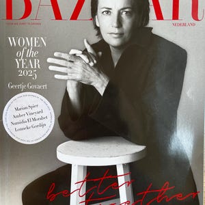 May include: A magazine cover featuring a woman in a black outfit, seated on a white stool. The magazine title "BAZAR" is in large red letters. Text includes "WOMEN of the YEAR 2025" and "Geertje Govaert". The words "Better Together" are written in red.