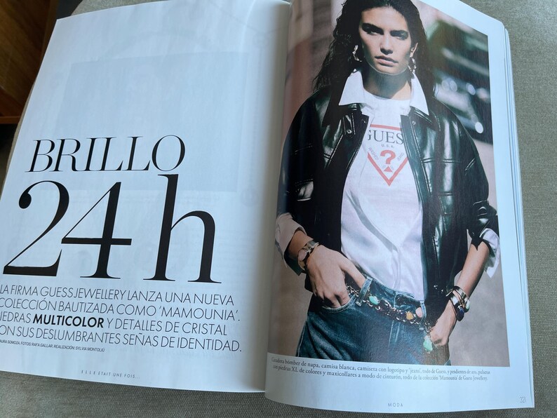Puede incluir: Una revista de moda con una modelo que lleva una chaqueta de cuero negra sobre una camiseta blanca de Guess y vaqueros. El texto "BRILLO 24h" aparece en la p&aacute;gina izquierda. La modelo tambi&eacute;n lleva joyas.