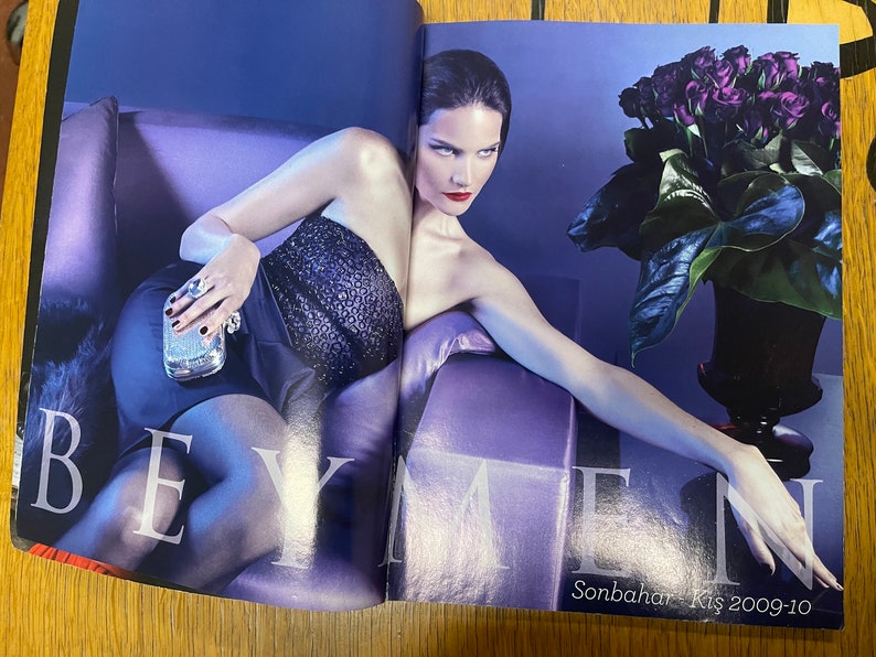 May include: A woman wearing a black sequined strapless dress and a silver clutch poses on a purple sofa. She has dark hair and red lipstick. A bouquet of dark purple roses in a black vase is in the background. The text "Sonbahar - Kis 2009-10" is visible in the bottom right corner.