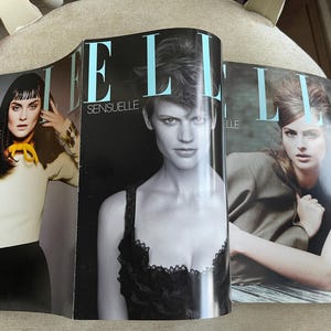 May include: Three open copies of Elle magazine are displayed, showcasing fashion photography. The covers feature models with various hairstyles and outfits. The magazine titles are visible, with the word "ELLE" in large, light blue letters. The cover text includes "INDIVIDUELLE" and "SENSUELLE".