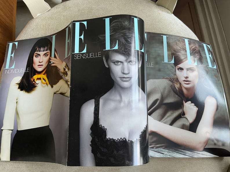 May include: Three open copies of Elle magazine are displayed, showcasing fashion photography. The covers feature models with various hairstyles and outfits. The magazine titles are visible, with the word "ELLE" in large, light blue letters. The cover text includes "INDIVIDUELLE" and "SENSUELLE".