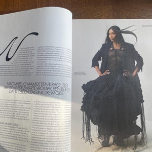 May include: A magazine spread showing a model in a black outfit. The ensemble includes a textured top, a long coat, and a skirt with fringe. The magazine page also features text in a foreign language.