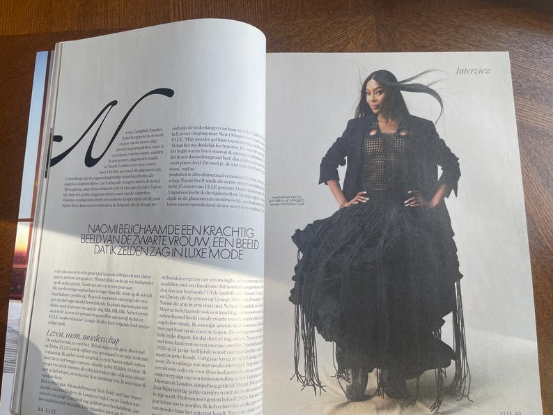 May include: A magazine spread showing a model in a black outfit. The ensemble includes a textured top, a long coat, and a skirt with fringe. The magazine page also features text in a foreign language.