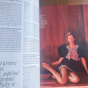 May include: A fashion editorial features a woman in a black outfit with a decorative, open-front jacket and a matching skirt. She is seated on a red rug against a reddish-brown wooden wall. The image is from a magazine.