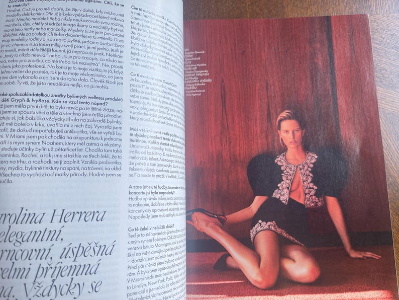 May include: A fashion editorial features a woman in a black outfit with a decorative, open-front jacket and a matching skirt. She is seated on a red rug against a reddish-brown wooden wall. The image is from a magazine.