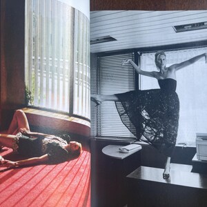May include: Black and white image of a woman in a strapless dress, striking a ballet pose on a desk. Another woman is lying on a red carpeted floor near a window with vertical blinds. The image is from a magazine.
