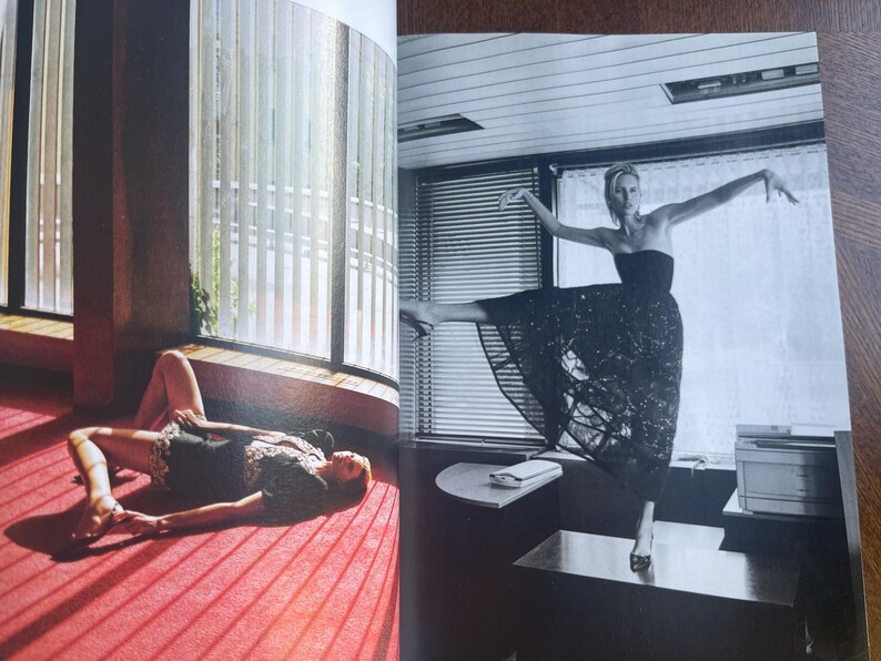 May include: Black and white image of a woman in a strapless dress, striking a ballet pose on a desk. Another woman is lying on a red carpeted floor near a window with vertical blinds. The image is from a magazine.