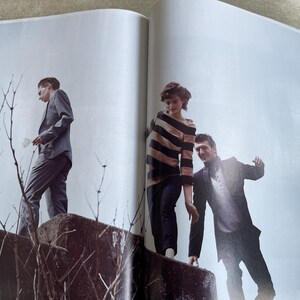 May include: Three people in stylish attire stand on a concrete structure against a light blue sky. One person wears a gray suit, another a striped sweater and dark pants, and the third a dark suit. The image is from a fashion magazine.