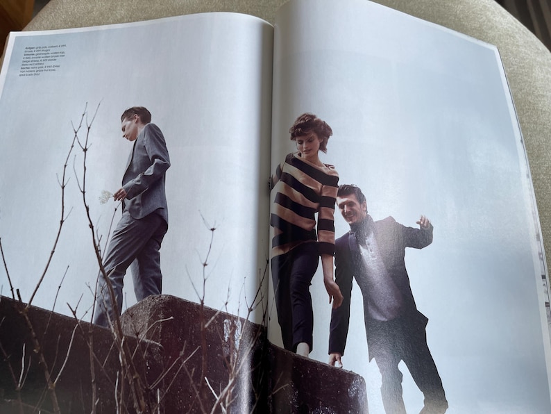 May include: Three people in stylish attire stand on a concrete structure against a light blue sky. One person wears a gray suit, another a striped sweater and dark pants, and the third a dark suit. The image is from a fashion magazine.
