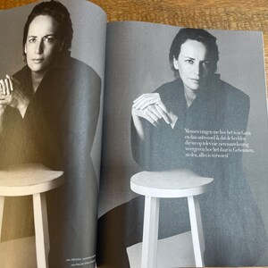 May include: A black and white magazine spread featuring a person in a dark outfit, seated on a small, white stool. The person is looking directly at the camera. Text in Dutch is visible on the right page.