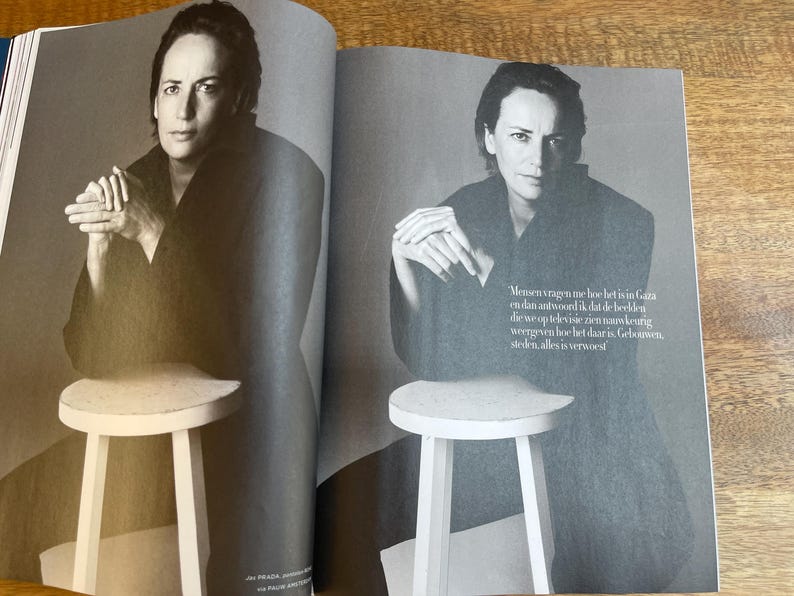 May include: A black and white magazine spread featuring a person in a dark outfit, seated on a small, white stool. The person is looking directly at the camera. Text in Dutch is visible on the right page.