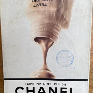 May include: An advertisement for Chanel Teint Naturel Fluide foundation. The image shows a bottle pouring out the product, with the brand name in bold black letters at the bottom. The background is a light, neutral color.