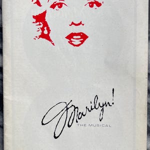May include: A vintage theater program cover featuring a stylized red illustration of Marilyn Monroe's face. The title "Marilyn! THE MUSICAL" is written in elegant script, with "ADELPHI THEATRE" below. The price is 50p.