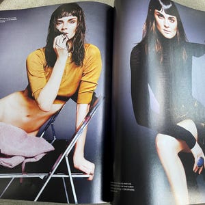 May include: A fashion magazine spread featuring two models. One model is seated in a mustard yellow long-sleeved top, the other in a black dress. The background is a gradient of gray.