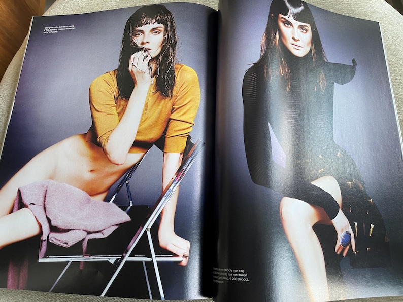 May include: A fashion magazine spread featuring two models. One model is seated in a mustard yellow long-sleeved top, the other in a black dress. The background is a gradient of gray.
