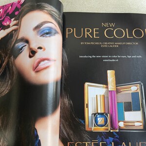 May include: An advertisement for Est&eacute;e Lauder's Pure Color makeup line. The image features a woman with blue eyeshadow and a variety of products, including a nail polish, lip gloss, and eyeshadow palette. The text reads "NEW PURE COLOR".