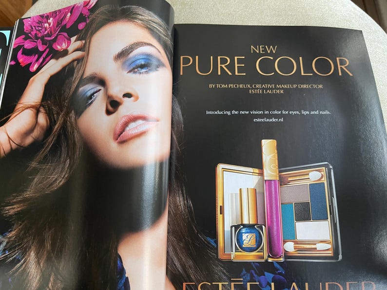 May include: An advertisement for Est&eacute;e Lauder's Pure Color makeup line. The image features a woman with blue eyeshadow and a variety of products, including a nail polish, lip gloss, and eyeshadow palette. The text reads "NEW PURE COLOR".