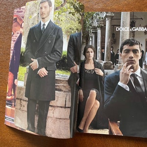 May include: A magazine spread featuring a fashion editorial with two models. The first model is wearing a black double-breasted coat and the second model is wearing a black suit with a white shirt and a black tie. The magazine cover features the text "DOLCE & GABBANA".