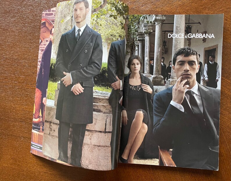 May include: A magazine spread featuring a fashion editorial with two models. The first model is wearing a black double-breasted coat and the second model is wearing a black suit with a white shirt and a black tie. The magazine cover features the text "DOLCE & GABBANA".